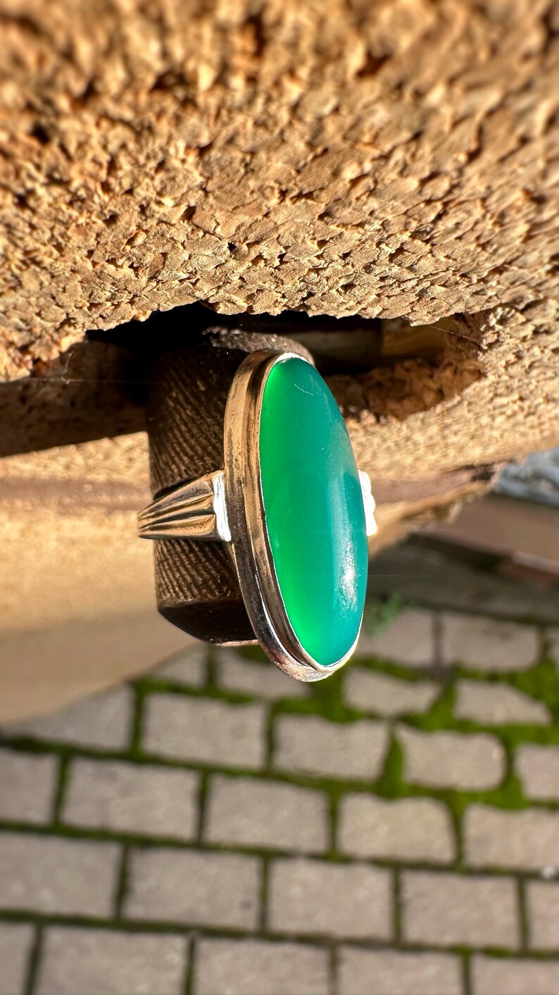 May include: A silver ring with a large, oval, bright green gemstone. The ring's band has a ribbed design on the sides. The gemstone is set in a silver bezel. The ring is displayed against a textured, brown background.