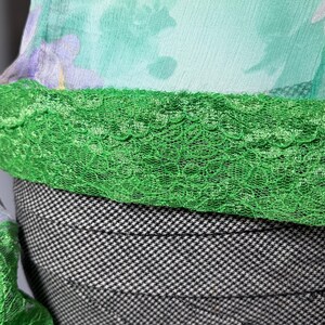 Op de afbeelding: Close-up van een vintage top met een bloemenprint in geel, paars en groen. De top heeft een groene kanten rand en wordt gecombineerd met een zwart-wit pied-de-poule rok. De stof lijkt licht van gewicht.