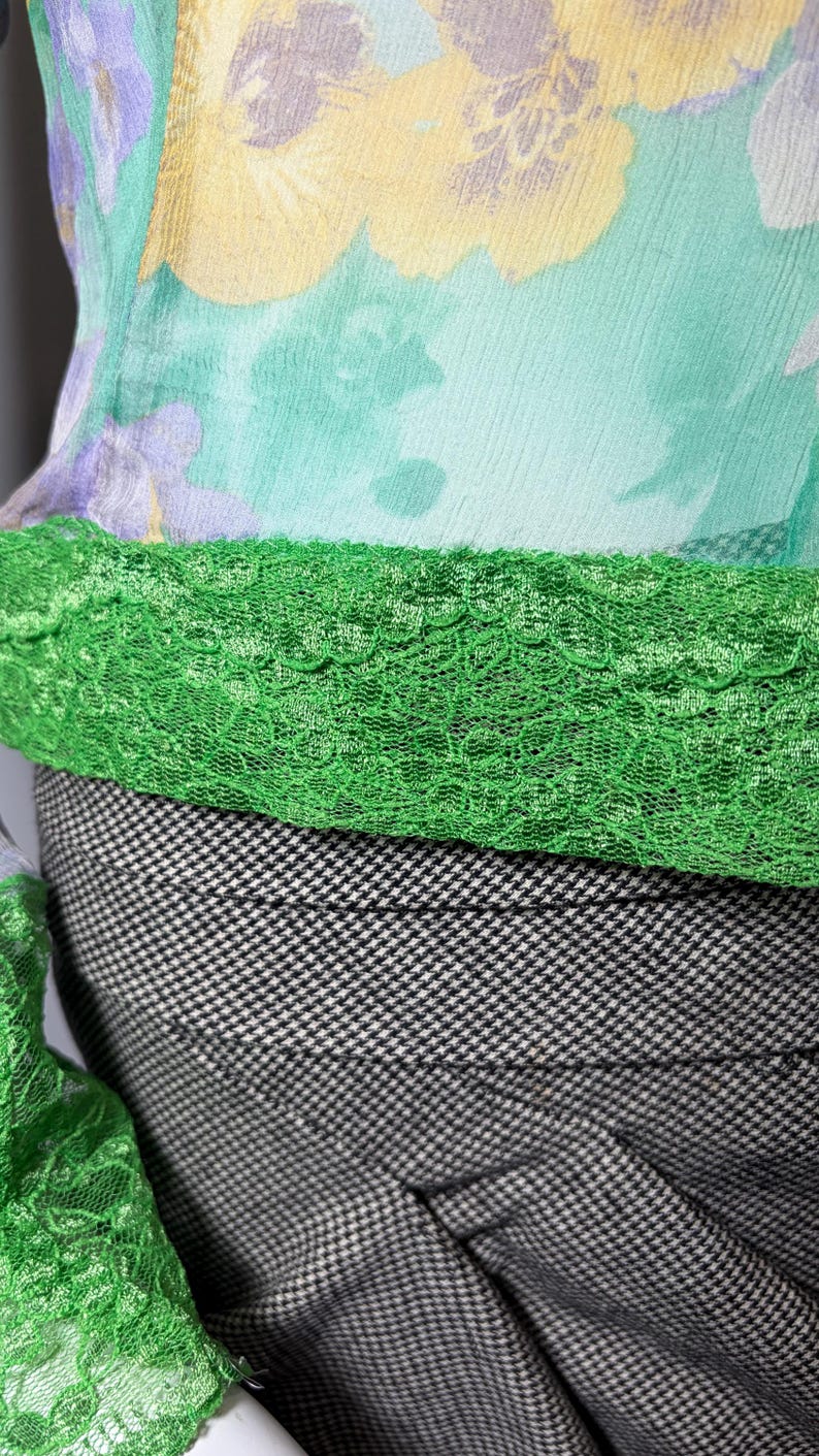 Op de afbeelding: Close-up van een vintage top met een bloemenprint in geel, paars en groen. De top heeft een groene kanten rand en wordt gecombineerd met een zwart-wit pied-de-poule rok. De stof lijkt licht van gewicht.