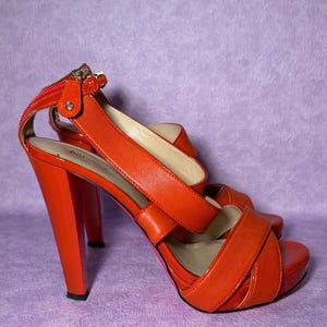 Peut inclure: Sandales à talons hauts orange vif avec un talon épais et une bride de cheville. Les sandales présentent un détail de fermeture éclair sur la bride de cheville et une semelle plateforme. Les chaussures sont en matière brillante, sur fond violet.