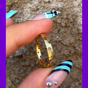 May include: A gold band ring featuring a series of small, circular, clear stones set at intervals along the outer surface. The metal has a polished finish, and the stones are embedded directly into the band, creating a continuous, repeating pattern.