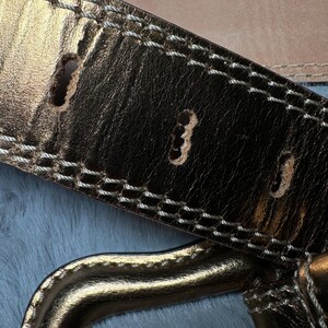 May include: Close-up of a gold-colored leather belt with white stitching and three belt holes. The belt has a metallic sheen and a gold-colored buckle. The background is a light blue, textured surface.