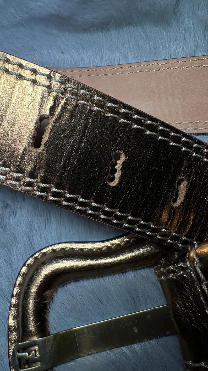 May include: Close-up of a gold-colored leather belt with white stitching and three belt holes. The belt has a metallic sheen and a gold-colored buckle. The background is a light blue, textured surface.