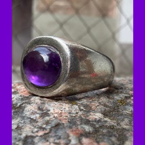 Puede incluir: Un anillo de metal plateado con una piedra preciosa de amatista de color púrpura intenso, lisa y redondeada, engastada en el centro. La banda tiene una construcción gruesa y sólida con un acabado pulido que muestra signos de desgaste y textura.