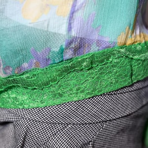 Op de afbeelding: Close-up van een top met bloemenpatroon en een groene kanten rand. De top heeft een mix van kleuren, waaronder geel, groen en paars. Onder de top zit een zwart-wit geruite stof.
