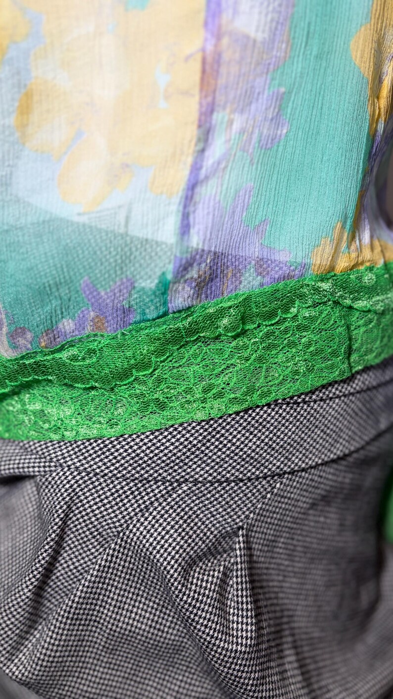Op de afbeelding: Close-up van een top met bloemenpatroon en een groene kanten rand. De top heeft een mix van kleuren, waaronder geel, groen en paars. Onder de top zit een zwart-wit geruite stof.