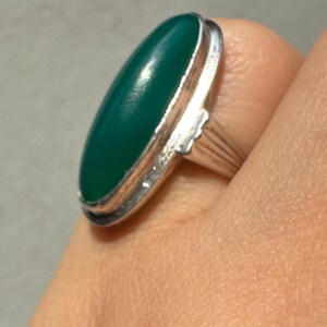 May include: A silver ring featuring a large, oval-shaped, emerald-green stone. The stone is set in a silver bezel and the ring band has a ribbed design. The ring is shown on a person's finger.