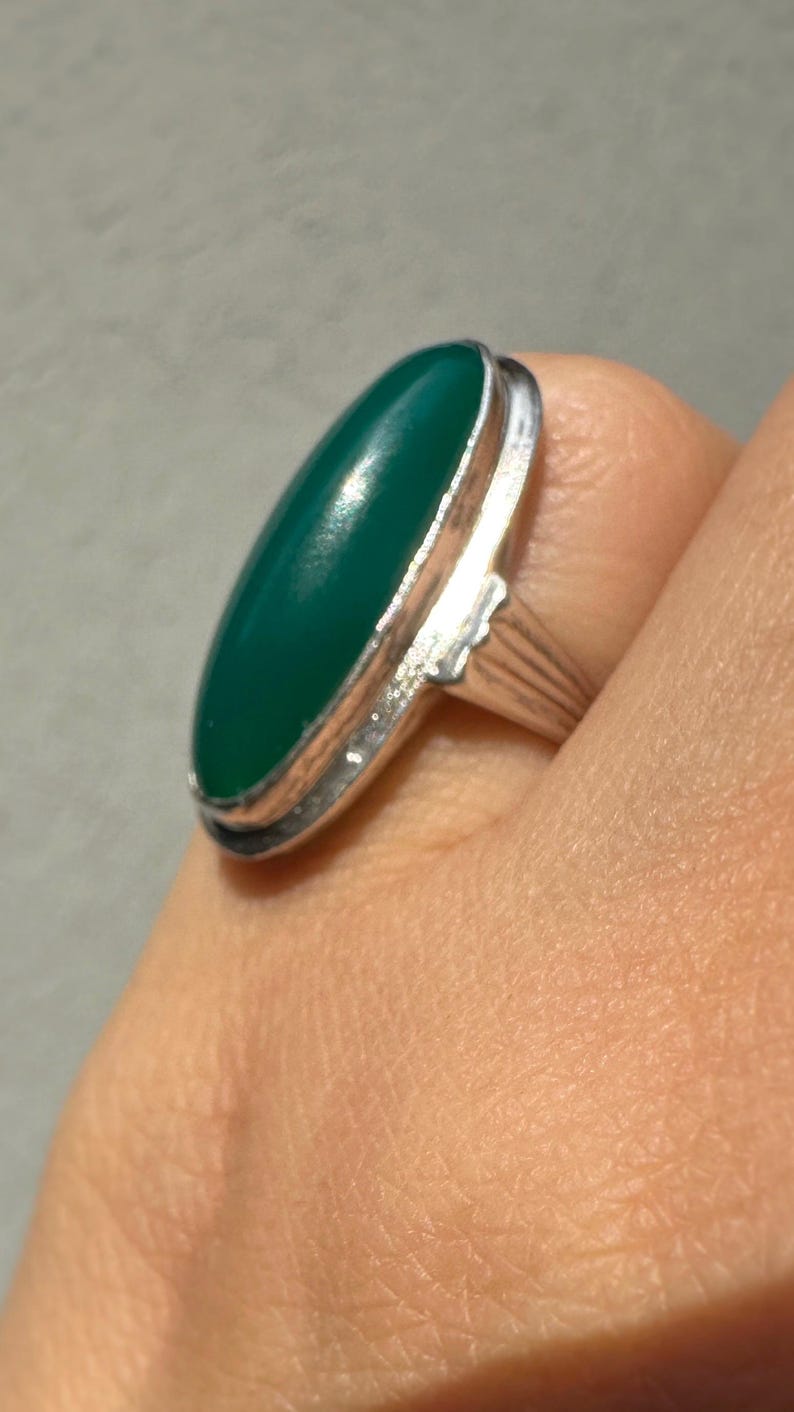 May include: A silver ring featuring a large, oval-shaped, emerald-green stone. The stone is set in a silver bezel and the ring band has a ribbed design. The ring is shown on a person's finger.