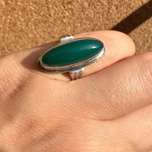 May include: A silver ring with an oval, deep green stone. The ring has a simple, elegant design with a polished finish. The stone is set in a bezel setting, and the band is smooth and unadorned. The ring is on a hand.