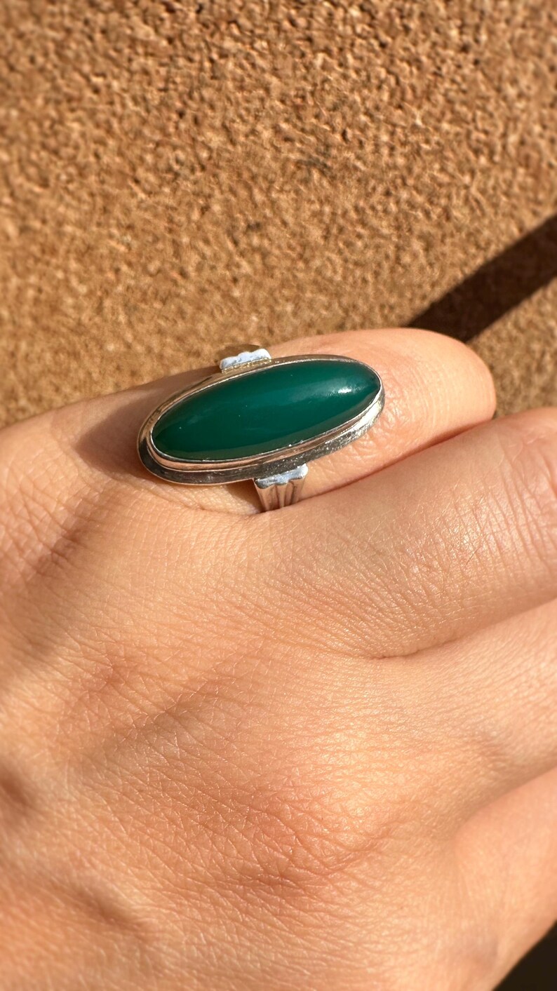 May include: A silver ring with an oval, deep green stone. The ring has a simple, elegant design with a polished finish. The stone is set in a bezel setting, and the band is smooth and unadorned. The ring is on a hand.
