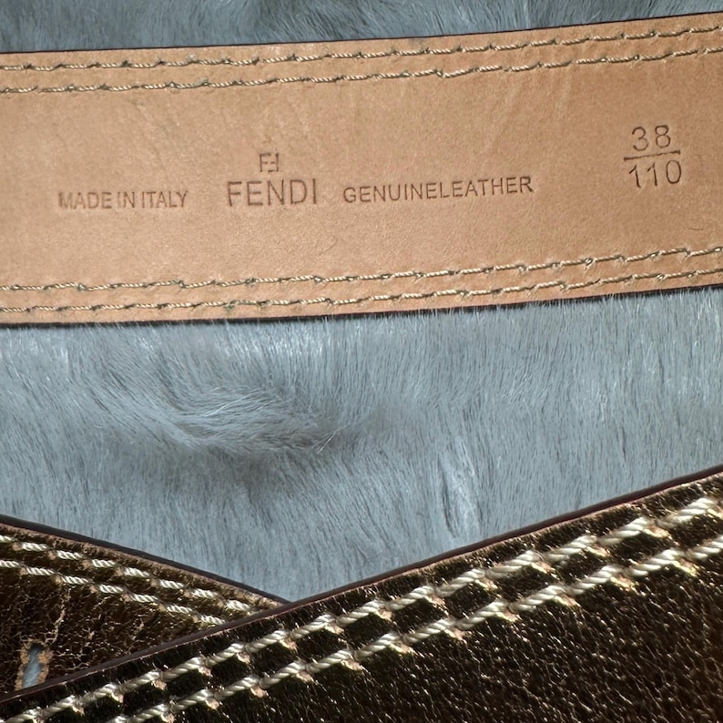 May include: Close-up of a Fendi genuine leather belt. The belt is brown with gold stitching and a gold metallic finish. The belt is marked "Made in Italy" and has the size 38/110.