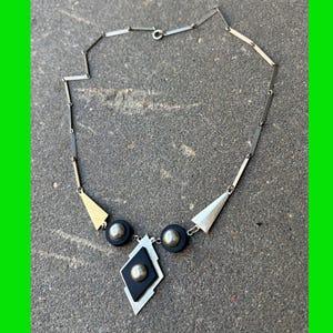 Puede incluir: Un collar de tono plateado con una cadena de eslabones rectangulares, dos acentos triangulares plateados, dos discos circulares negros con esferas plateadas centrales y un colgante negro en forma de diamante con una esfera plateada.