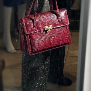 Könnte beinhalten: Eine weinrote Handtasche mit einem strukturierten, schlangenlederartigen Muster. Die Tasche hat eine rechteckige Form mit einem Klappenverschluss und einem goldfarbenen Verschluss. Sie hat zwei Griffe und eine Vordertasche.