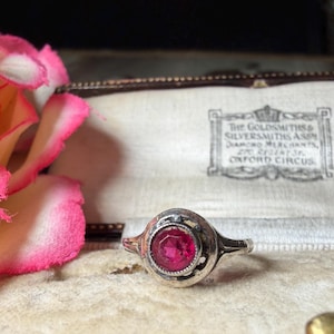 May include: A silver ring featuring a round, faceted, deep pink gemstone set in a circular bezel. The ring is displayed inside a vintage jewelry box with a cream-colored interior lining that includes the printed text: The Goldsmiths & Silversmiths Assn.