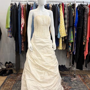 May include: A cream-colored strapless wedding dress on a mannequin. The bodice is embellished with small, sparkling details, and the skirt features draped fabric. The dress is displayed in a vintage clothing store, with racks of clothing and posters in the background.