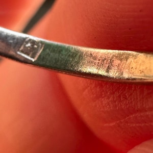 May include: Close-up of a silver ring with a small, square hallmark. The ring's surface shows wear and tear, with visible scratches. The background is a blurred view of skin.