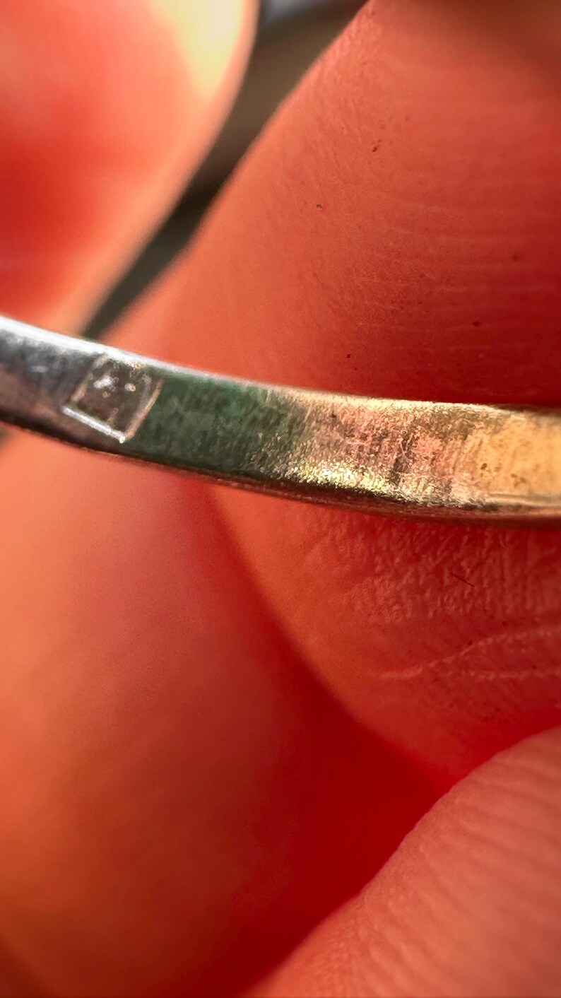 May include: Close-up of a silver ring with a small, square hallmark. The ring's surface shows wear and tear, with visible scratches. The background is a blurred view of skin.