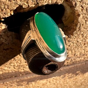 May include: A silver ring with a large, oval, bright green stone. The ring has a simple, elegant design with a polished finish. The gemstone is the focal point, set in a bezel setting. The ring is displayed against a textured background.