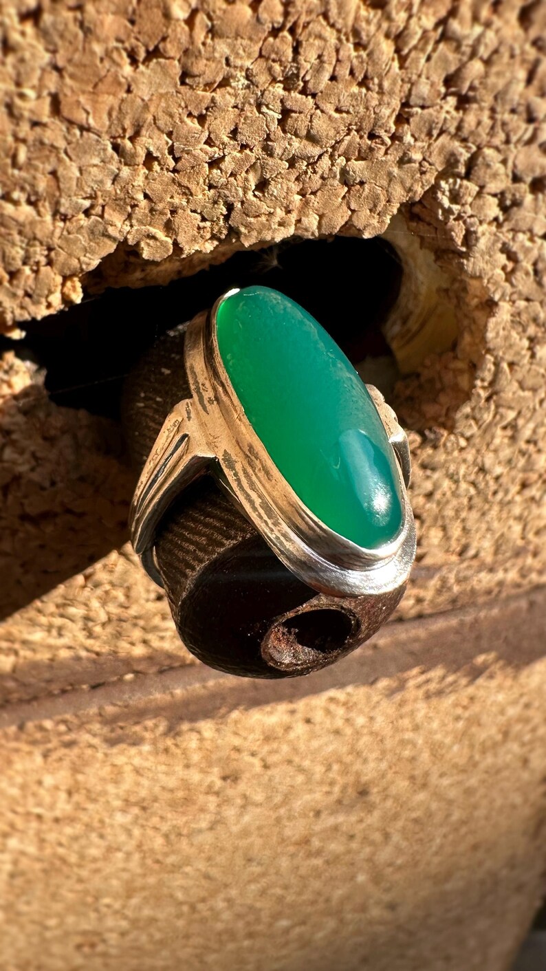 May include: A silver ring with a large, oval, bright green stone. The ring has a simple, elegant design with a polished finish. The gemstone is the focal point, set in a bezel setting. The ring is displayed against a textured background.