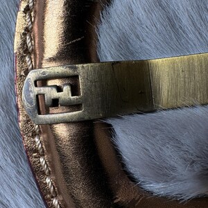 May include: Close-up of a gold-toned metal clasp with a geometric design, attached to a metallic copper-colored leather strap. The strap is stitched with a light-colored thread. The background is a soft, white, furry texture.