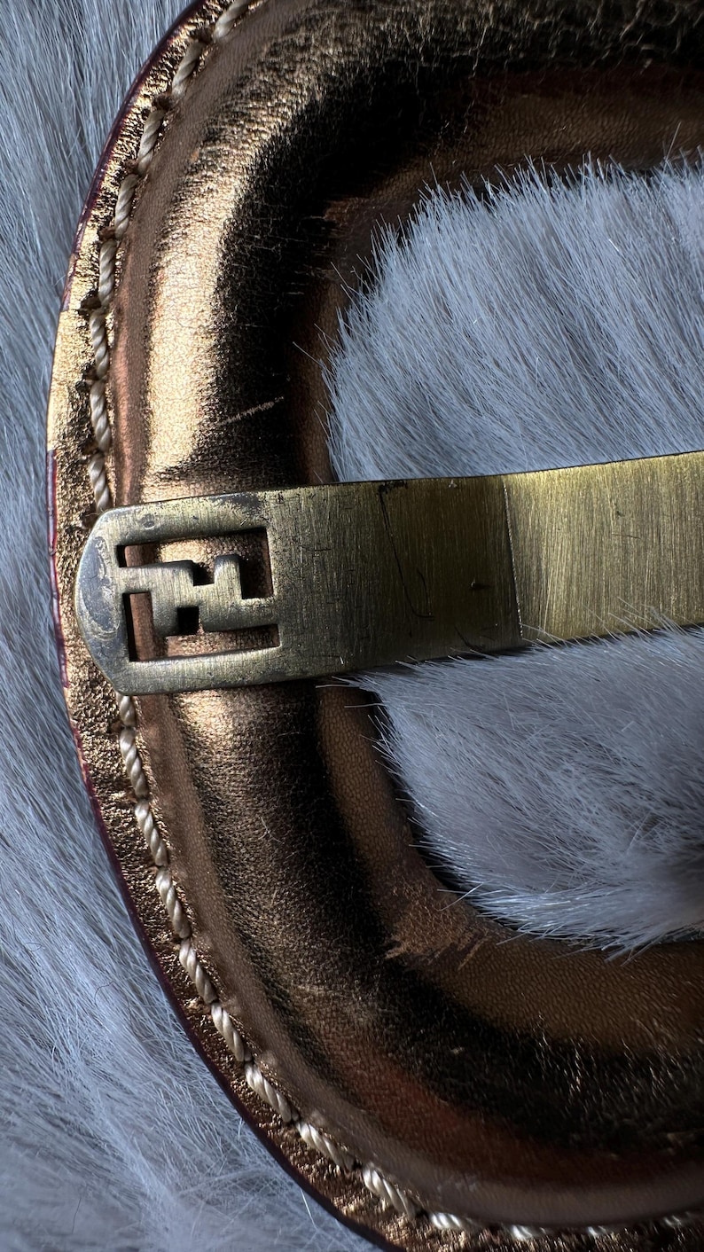 May include: Close-up of a gold-toned metal clasp with a geometric design, attached to a metallic copper-colored leather strap. The strap is stitched with a light-colored thread. The background is a soft, white, furry texture.
