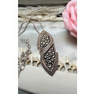 Op de afbeelding: Zilverkleurige ketting met een hanger met een gedetailleerd, verweven ontwerp. De hanger is bezet met kleine, donkere stenen, wat een sprankelend effect creëert. De ketting wordt gepresenteerd op een lichtgekleurd oppervlak, met een roze bloem op de achtergrond.
