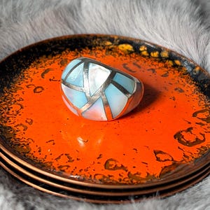 May include: A silver ring with a geometric design featuring light blue and white inlays, resting on a stack of orange plates with a black and gold rim. The plates have a textured surface.