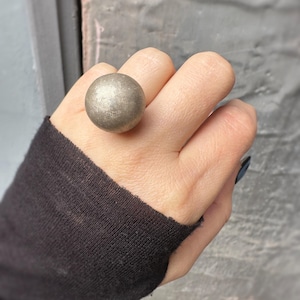May include: A silver-colored, spherical ring is worn on a finger. The ring's large, round design is the focal point, with a textured surface. The person is wearing a black long-sleeved shirt.