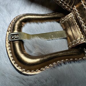 May include: Close-up of a gold-colored belt with a rectangular buckle. The belt has a stitched design along the edges and multiple holes for adjustment. The buckle features a logo. The belt is set against a white, furry background.