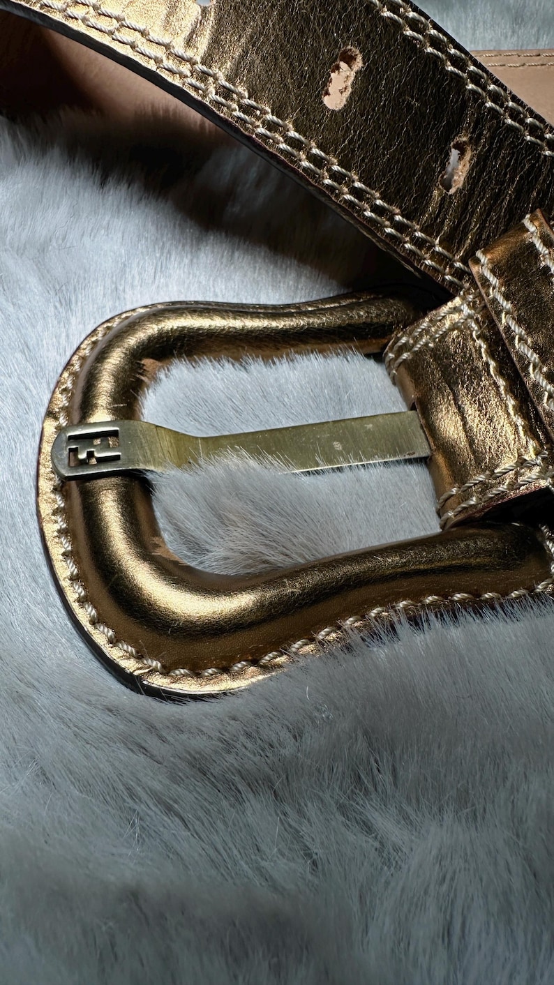 May include: Close-up of a gold-colored belt with a rectangular buckle. The belt has a stitched design along the edges and multiple holes for adjustment. The buckle features a logo. The belt is set against a white, furry background.