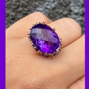 May include: A ring featuring a large, oval-cut, deep purple amethyst gemstone with a faceted surface. The stone is secured in a metallic setting with multiple prongs that extend around the perimeter of the gem, creating a crown-like appearance.