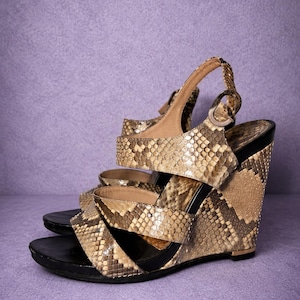 May include: A pair of wedge sandals with a snakeskin pattern in shades of tan, brown, and black. The sandals have a black sole and straps that wrap around the foot and ankle. The background is a solid purple.