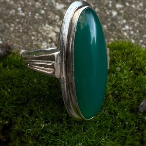 May include: A silver ring with a large, oval, emerald-green stone. The ring has a simple, elegant design with a polished finish. The stone is set in a bezel setting, and the band has a decorative design on the sides.