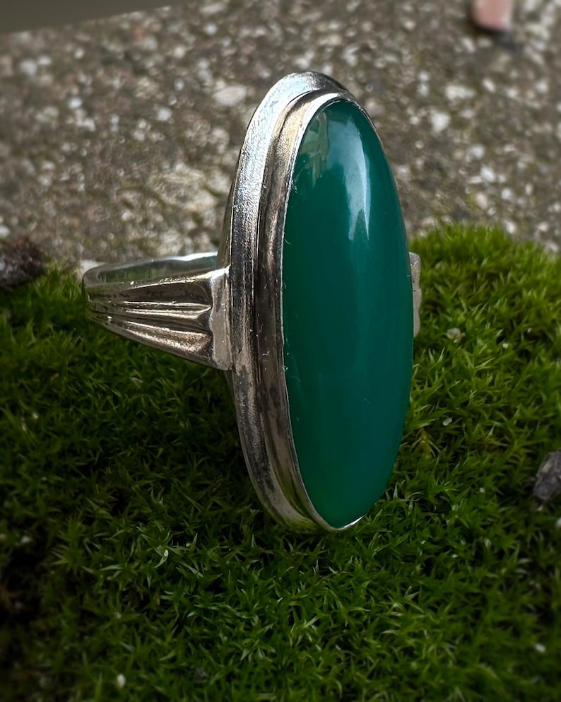 May include: A silver ring with a large, oval, emerald-green stone. The ring has a simple, elegant design with a polished finish. The stone is set in a bezel setting, and the band has a decorative design on the sides.
