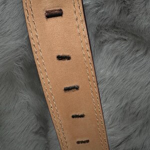 May include: Close-up of a tan leather belt with evenly spaced rectangular holes. The belt has a stitched border with a lighter thread. The background is a soft, gray, furry texture.