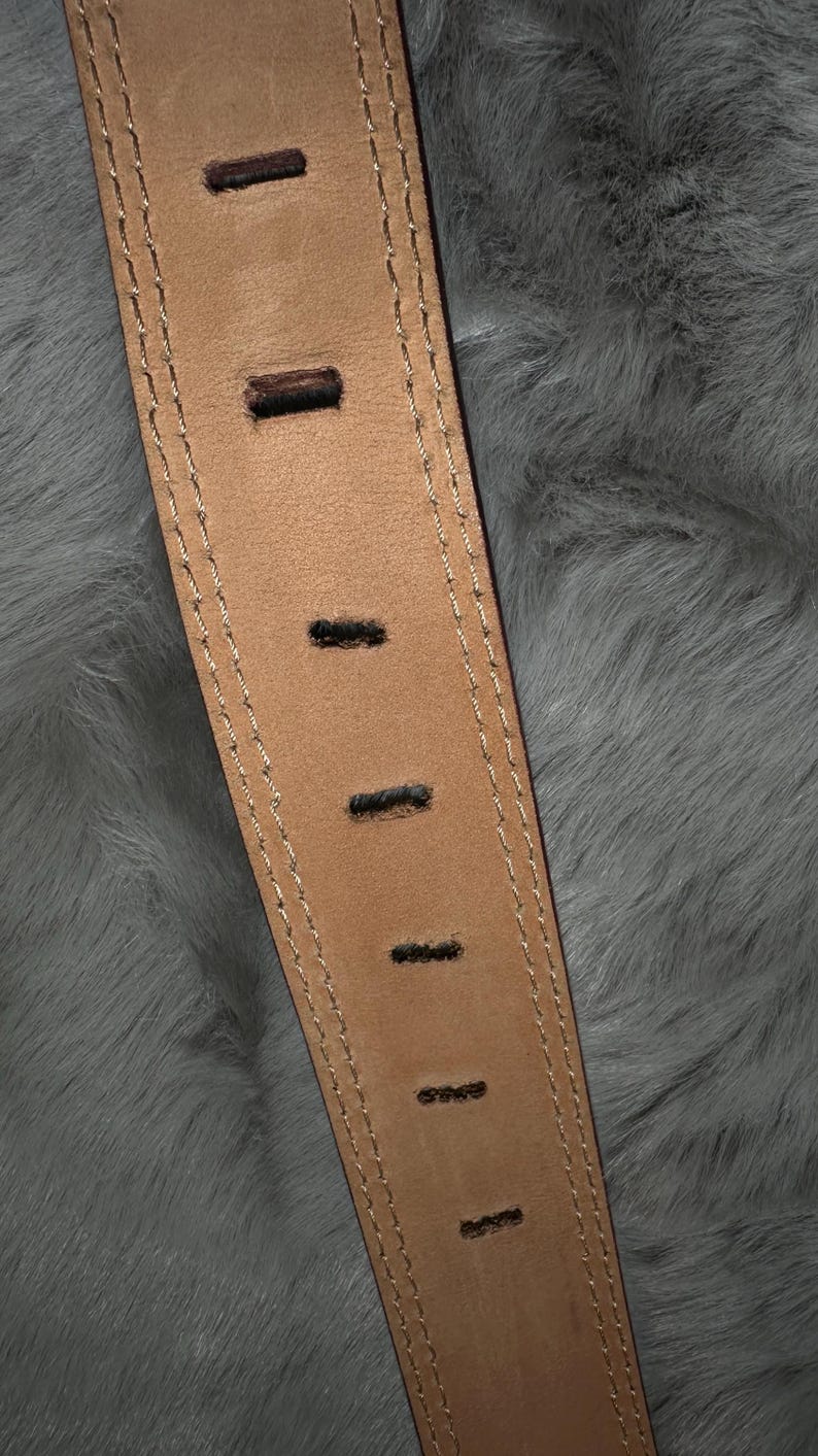 May include: Close-up of a tan leather belt with evenly spaced rectangular holes. The belt has a stitched border with a lighter thread. The background is a soft, gray, furry texture.