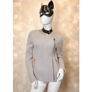 May include: A mannequin wearing a light grey ribbed knit jacket with an asymmetrical zip. The mannequin also wears a black leather cat-ear mask and a black choker. The background is a soft, blurred pattern of light.
