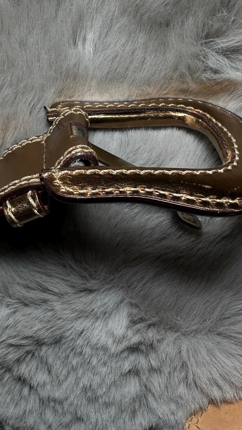 May include: Close-up of a brown leather handle with gold accents and white stitching. The handle is set against a backdrop of gray faux fur, creating a textural contrast. The handle appears to be part of a bag or purse.