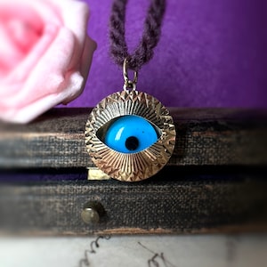 May include: A silver necklace with a blue evil eye charm. The charm is surrounded by a gold-colored, textured frame. The necklace is on a dark wooden surface.