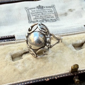 May include: A silver ring featuring a central polished sphere surrounded by an openwork frame with leaf and scroll motifs. The ring is presented in a vintage jewelry box with a label reading The Goldsmiths & Silversmiths Assn Diamond Merchants 270 Regent St Oxford Circus.