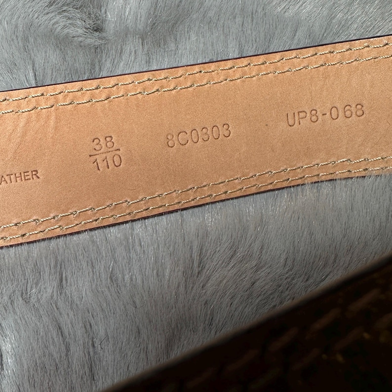 May include: Close-up of a brown leather belt's interior. The belt is stitched with a light-colored thread. The size is indicated as 38 inches and 110 cm. The belt also has the numbers 8C0303 and UP8-068 printed on it.