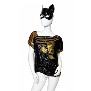 Op de afbeelding: Een top met korte mouwen bedekt met zwarte en gouden lovertjes, voorzien van een wijde halslijn en een geometrisch patroon. Het kledingstuk wordt getoond op een witte etalagepop die een zwart masker met kattenoren over het gezicht draagt.