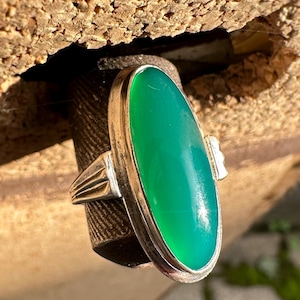 May include: A silver ring with a large, oval, bright green gemstone. The ring has a simple, elegant design with a rectangular accent on the side. The gemstone is set in a silver bezel, and the ring is displayed against a textured background.