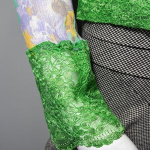 Op de afbeelding: Een mouw met bloemenprint en groene kanten afwerking. De mouw heeft een patroon van gele, paarse en lichtblauwe bloemen. De manchet en halslijn zijn afgezet met heldergroen kant.