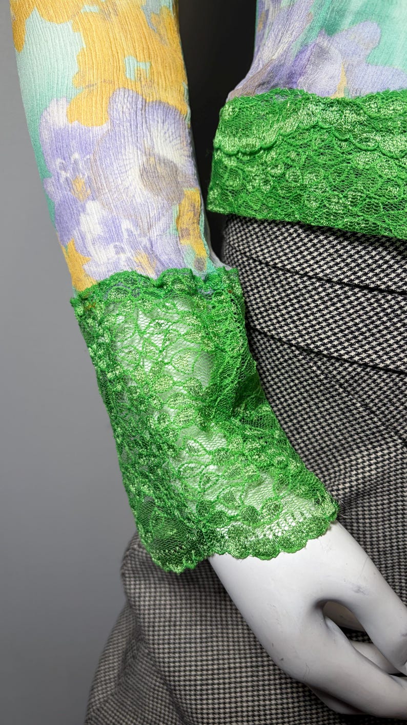 Op de afbeelding: Een mouw met bloemenprint en groene kanten afwerking. De mouw heeft een patroon van gele, paarse en lichtblauwe bloemen. De manchet en halslijn zijn afgezet met heldergroen kant.