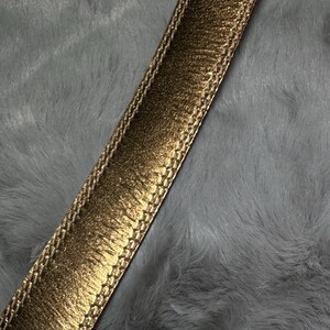 May include: Close-up of a gold-colored belt with a textured, metallic finish. The belt has a stitched border and is set against a gray, fluffy background. The belt's design suggests a fashion accessory.