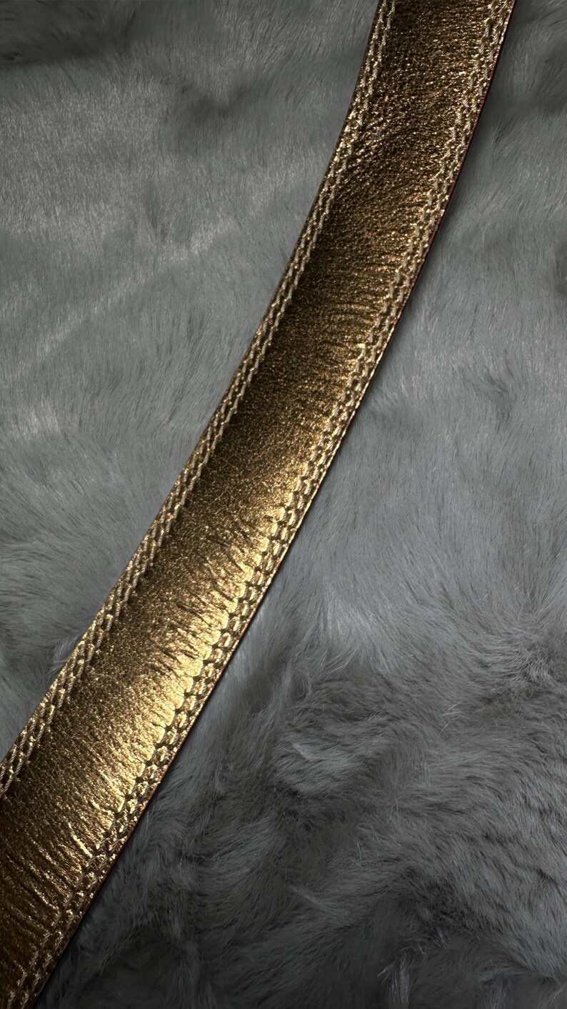May include: Close-up of a gold-colored belt with a textured, metallic finish. The belt has a stitched border and is set against a gray, fluffy background. The belt's design suggests a fashion accessory.