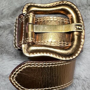 May include: A gold-colored belt with a rectangular buckle and a matching belt loop. The belt is made of a shiny, metallic material and has a stitched edge. The buckle has a simple, classic design.