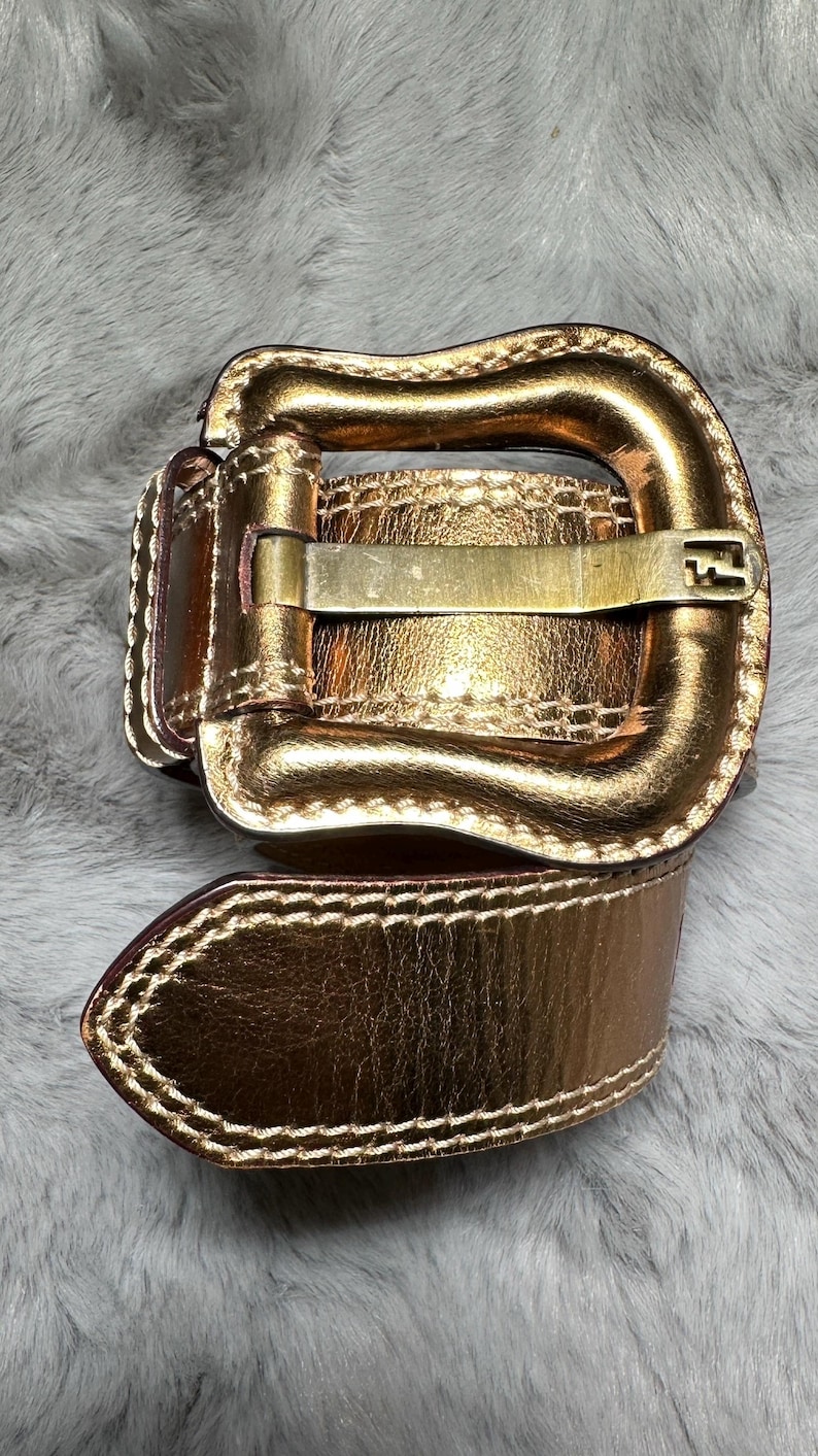 May include: A gold-colored belt with a rectangular buckle and a matching belt loop. The belt is made of a shiny, metallic material and has a stitched edge. The buckle has a simple, classic design.