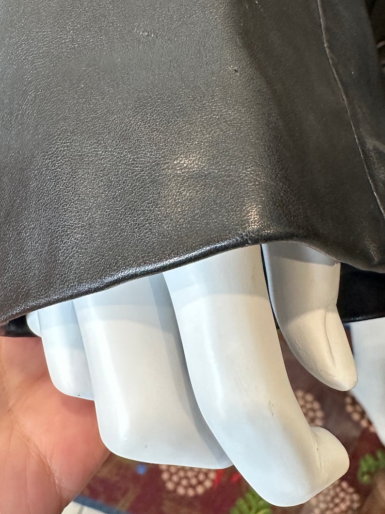 May include: Close-up of a black leather jacket sleeve with a textured surface. The sleeve is folded over a white mannequin hand.